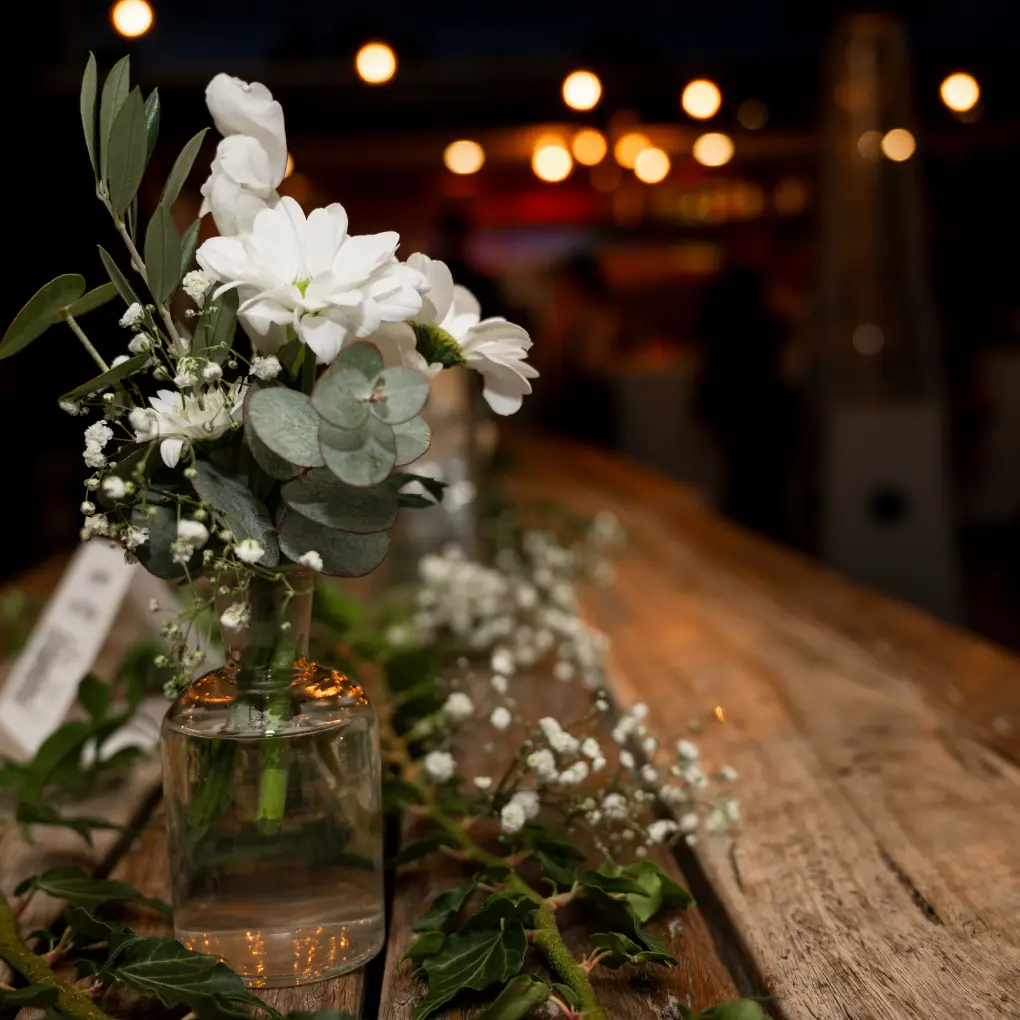 birthday-party-flower-decor Elegant white floral arrangement in a glass vase styled on a rustic timber table for an event or wedding setting with warm ambient lighting.