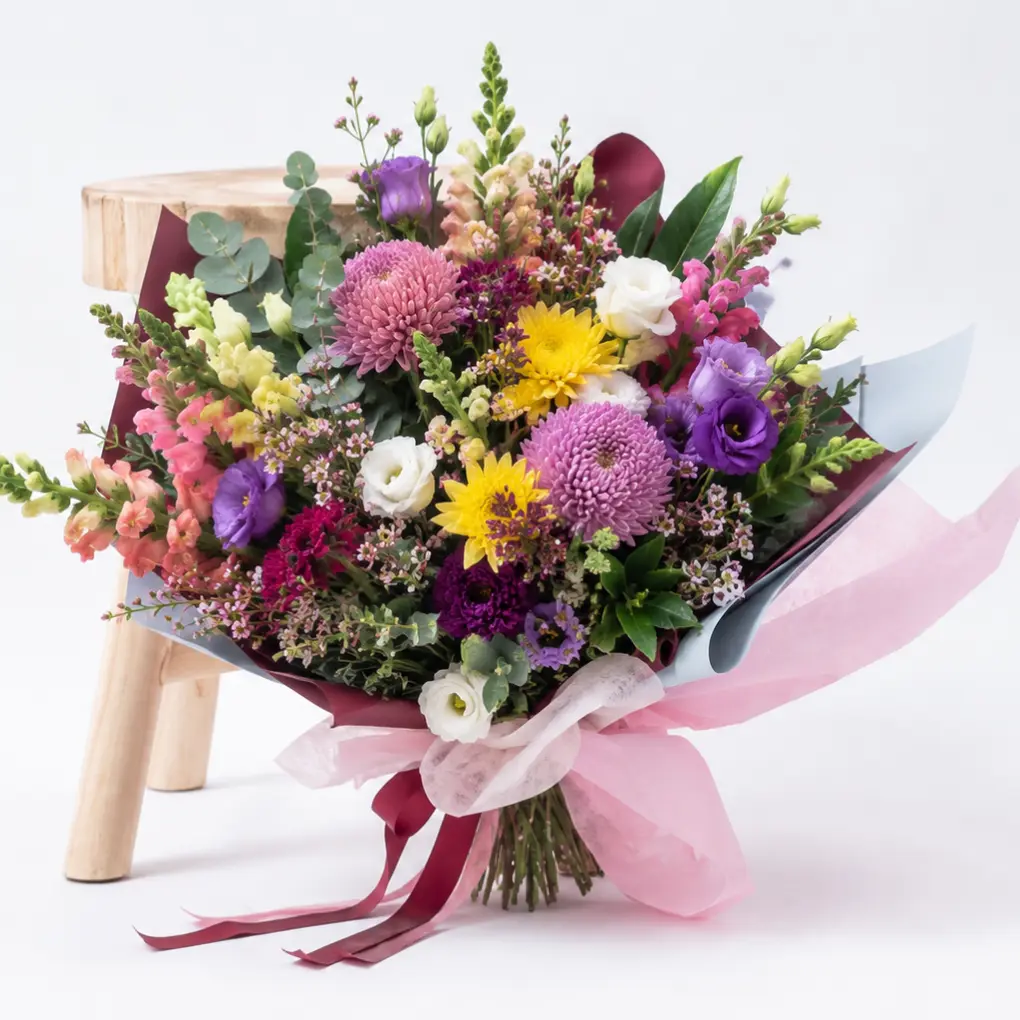 corporate-flowers Colourful everyday flower bouquet with chrysanthemums, lisianthus, snapdragons and wax flowers styled beside a wooden stool.