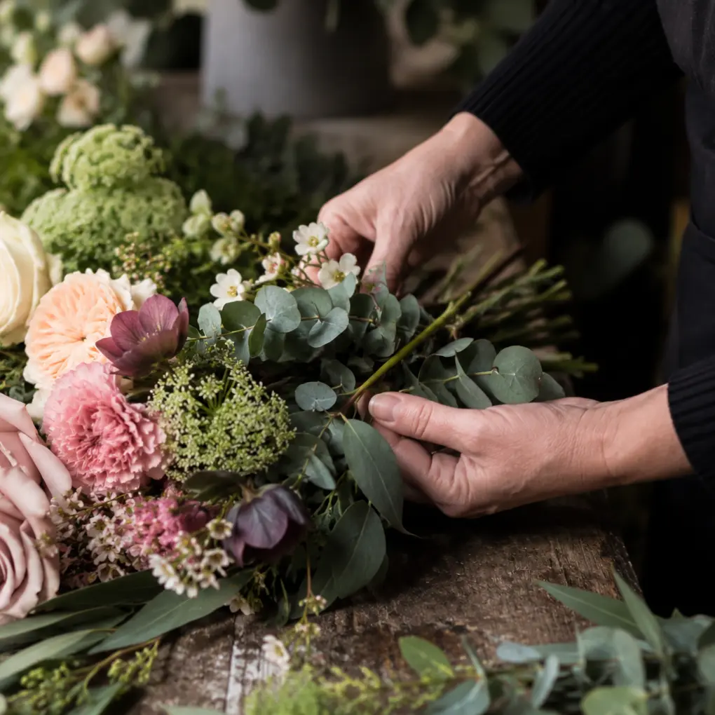 Florist arranging soft seasonal flowers and greenery on a rustic workbench with pastel roses and eucalyptus.